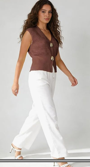 Woman wearing a brown sleeveless top with buttons and white pants on a plain background