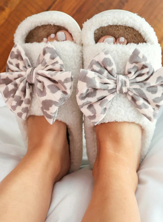 White slippers with leopard print bows worn by a person on a wooden floor.