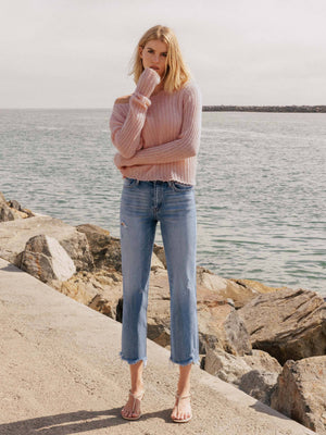 Woman wearing a pink sweater and blue jeans standing on rocks by the water.